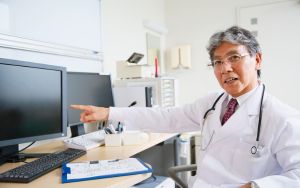 Bác sĩ lớn tuổi mặc áo blouse và đeo ống nghe đang ngồi tại bàn làm việc, chỉ tay vào màn hình máy tính để giới thiệu các phương pháp kiểm soát cận thị mới nhất 