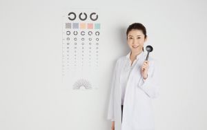 Bác sĩ nhãn khoa cầm dụng cụ kiểm tra mắt đứng cạnh bảng đo thị lực trong phòng khám trắng để giải thích về tầm quan trọng của kiểm soát cận thị